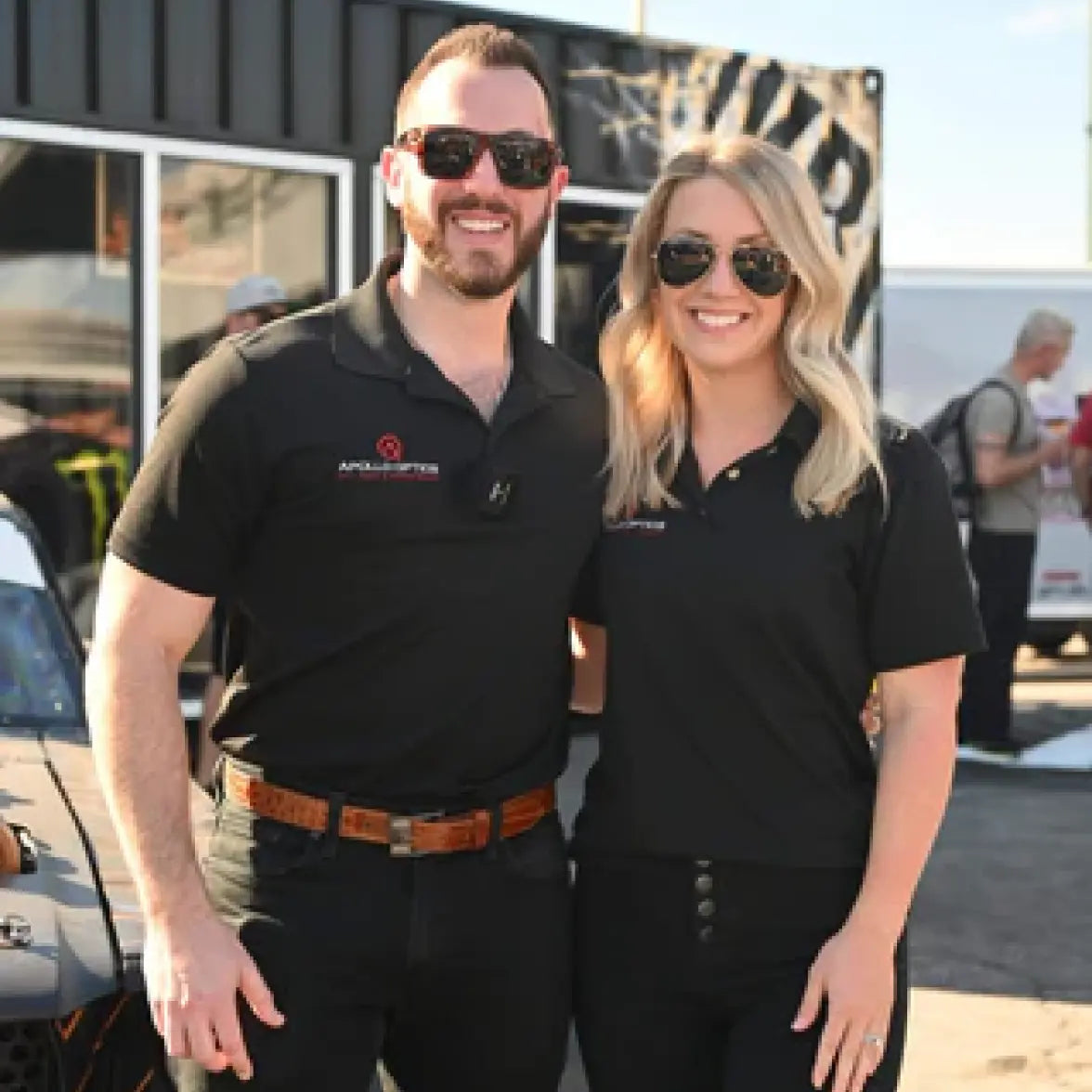 A man and a woman standing side-by-side in matching black polo shirts with the 'Applause' brand logo on the chest, wearing sunglasses and posing outdoors in front of a building.